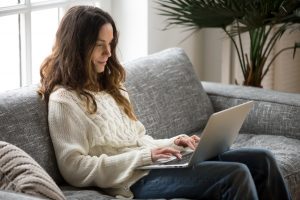 A woman smiles as she sits on her couch and types on her laptop. This could represent the positives of online therapy in Florida. Contact an online therapist in Florida for support with online EMDR therapy and other services. 