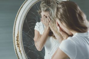 A woman covers her face while standing next to a cracked mirror. Learn how a DBT therpaist in St. Petersburg, FL can offer support in overcoming emotional distress. Learn more about DBT therapy in St. Pete, FL.