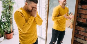 A person covers their face while standing next to a mirror with a laughing reflection. A DBT therapist in St. Petersburg, FL can offer emotional regulation support. Learn more about dialectical behavior therapy in St. Pete, FL to learn more about DBT today.