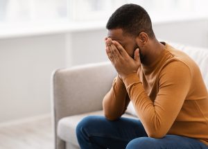A man covers his face while sitting on the end of a sofa. Learn more about the support a therapist in St. Pete, FL can offer by searching for a St. Pete therapist or Tampa therapist today.