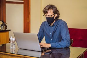 A man wears a mask while typing on a laptop. Learn how online therapy in Florida can offer support by contacting an online therapist in Florida. They can offer support with online couples counseling in Florida and other services.