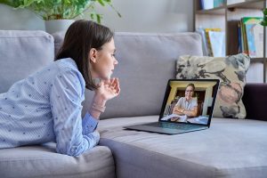 A woman lays on her stomach while talking on a zoom call on the couch. Learn how Psypact in Florida can offer support with the help of psypact therapists in florida. Learn more about psypact therapists in south carolina and other states like Alabama.